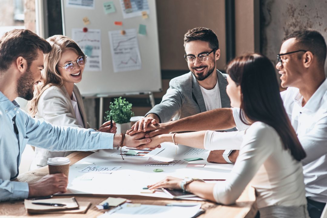 A group of five people sitting around a table working on a project with their all their hands connected in the middle