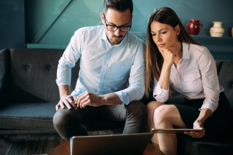 Man and woman working on laptop and tablet. Woman has long hair and is leaning on hear knee